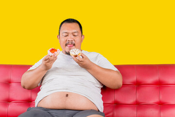 Overweight Asian guy sitting on the sofa while eating delicious donuts