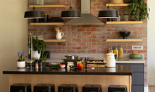 Domestic kitchen with shelves and hood over hob on exposed brick wall and hanging lamps over island