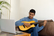 © WavebreakMediaMicro - Biracial man sitting on sofa playing guitar and using laptop at home