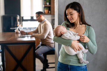 Wall Mural - Young father works on laptop while his wife looks after an infant child. Work from home