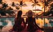 Rear back view two women in hat and summer dress welcoming sunset seated on poolside, enjoy evening light and view to exotic tropical nature on holidays in luxury resort. Vacation, travel concept