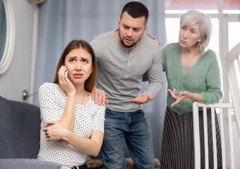 Wall Mural - Sad young woman sitting at home ignoring her husband and mother-in -law berating her