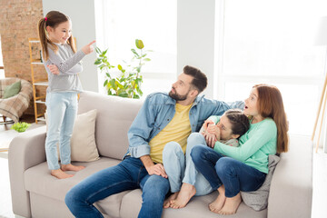 Wall Mural - Photo of four persons sit on couch small positive girl standing point finger tell story to daddy astonished sisters indoors