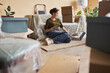 © Seventyfour - Young woman with smartphone in hand typing on laptop keyboard while sitting on the floor by couch covered with cellophane after relocation