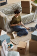 © Seventyfour - Young brunette female freelancer using laptop while sitting on the floor of living room with packed boxes after relocation to new apartment