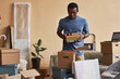 © Seventyfour - Young African American man with stack of books packing or unpacking boxes while standing in living room of new apartment or house