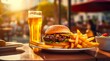 Tasty hamburger, french fries and glass of light beer on the table on the blurred background of restaurant outdoor at sunny day.