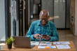 A black senior man in casual clothes uses a cell phone to find information or talk on a touch screen while working at a desk.
