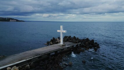 Wall Mural - A large Christian cross stands at the edge of a pier against a dramatic sky and sea, seen from above