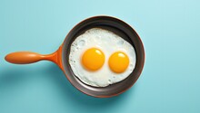  Two Fried Eggs In A Frying Pan On A Blue Background With A Wooden Spoon In The Middle Of The Frying Pan.