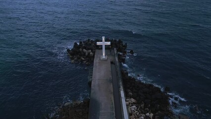 Wall Mural - A large Christian cross stands at the edge of a pier against a dramatic sky and sea, seen from above