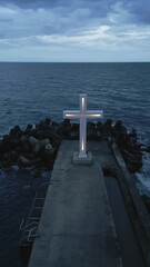 Wall Mural - A large Christian cross stands at the edge of a pier against a dramatic sky and sea, seen from above