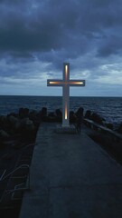 Wall Mural - A large Christian cross stands at the edge of a pier against a dramatic sky and sea, seen from above