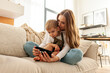 young mother and her son are sitting on the sofa and using smartphone, woman is hugging child and looking at the phone