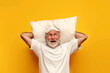 old bald grandfather in pajamas and sleep mask lies on pillow and imagines on yellow isolated background
