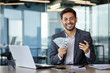 © Liubomir - Arab man in a business suit holds a phone and a stack of dollar bills in his hands, sits at a desk in the office, rejoices at the win, smiles and looks at the camera.