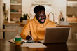A cheerful interracial man is writing down notes while smiling at the laptop late at night from home.
