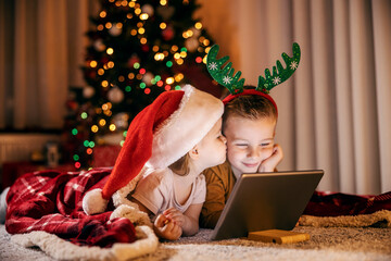 Little brother and sister kissing and watching videos on tablet on new year and christmas eve.