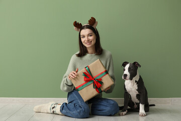 Wall Mural - Beautiful young woman in reindeer horns with Christmas gift and cute Staffordshire Terrier puppy near green wall