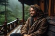 stylish man traveler in blanket relaxing on porch of wooden cabin in rainy day on background of woods