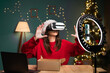 © Andrii Lysenko - blogging, technology and people - happy smiling girl blogger in santa hat with ring light and smartphone doing VR glasses test live. concept of advertising products to bloggers on Christmas Eve