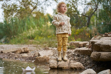 Smiling Girl Standing On Rock With Paper Boat In Water Puddle