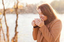 Smiling woman drinking tea from cup by lake at sunset