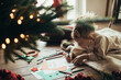 © polinaloves - Cute girl writing a letter to Santa Claus, lying on a wooden floor under decorated Christmas tree.