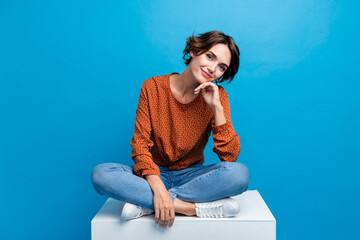 Full length photo of lovely young lady sit cube look dreamy camera dressed stylish brown garment isolated on blue color background