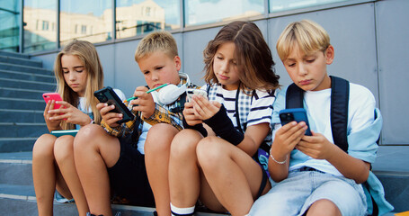 Tenager schoolchildren of two Caucasian girls and boys holding mobile phone and browsing while sitting on stair during school break, oudoors