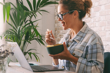 Wall Mural - Side view of adult young woman smiling and eating in fron tof a laptop at the desk at home. Alternative office lifestyle female people working in video call conference during lunch time. Modern job