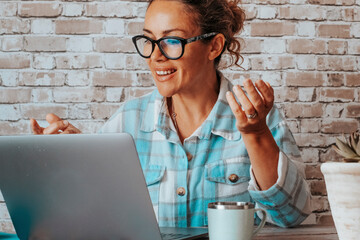 Wall Mural - Hipter young woman talking in video call conference onine on laptop. People at work with computer. Bricks background and happy lady speaking and enjoying communication modern job