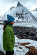 Lofoten islands, Senja region, Norway. Winter. Young girl in green jacket and blue hat stands on ocean. Wind blows. In background snowy mountains. Concept of tourism
