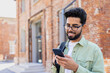 © Liubomir - Portrait of young successful student outside university campus, man smiling and using app on phone, typing message and browsing social media, indian man holding phone with backpack