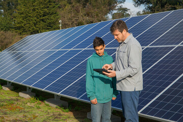 Father and adolescent son checking the efficiency of solar panels with a digital tablet, promoting eco-friendly initiatives.