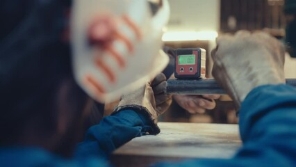 Metalworker in protective gloves and welding helmet using digital level box displaying angle of metal pipe surface at workbench in repair shop. Over-the-shoulder shot, selective focus