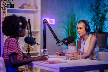 Wall Mural - Two interracial co-hosts are sitting in a home broadcast studio with headphones on and discussing topics while using microphones.