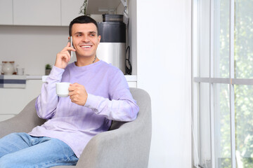 Poster - Handsome young man drinking coffee while talking by mobile phone and sitting on armchair near window