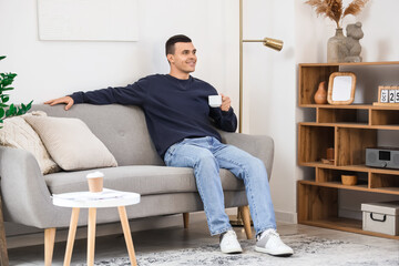 Poster - Handsome young man with cup of coffee sitting on sofa in modern living room