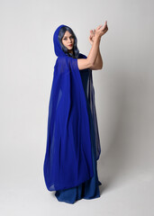 Poster -  Full length portrait of beautiful female model wearing elegant fantasy blue ball gown, flowing cape with hood.
Standing pose walking away, gestural arms reaching out. Isolated on  studio background