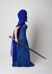 Canvas Print - Full length portrait of beautiful female model wearing elegant fantasy blue ball gown, flowing cape with hood.
Standing pose walking away, holding a sword weapon Isolated on white studio background.