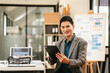 © Phushutter - Attractive professional Asian businessman with tablet in office environment, confidently smiling, possibly analyzing data as Capital Market Analyst.