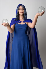 Wall Mural - Close up portrait of beautiful female model wearing elegant fantasy blue ball gown, flowing cape with hooded cloak.
 Holding a crystal glass seer orb. Isolated on white studio background.