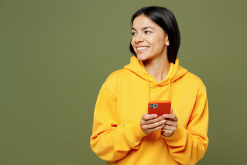 Wall Mural - Young smiling happy Latin woman she wear yellow hoody casual clothes hold in hand use mobile cell phone look aside on area isolated on plain pastel green background studio portrait. Lifestyle concept