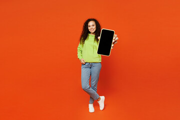 Wall Mural - Full body young fun woman of African American ethnicity she wear green hoody casual clothes hold in hand use mobile cell phone with blank screen workspace area isolated on plain red orange background.
