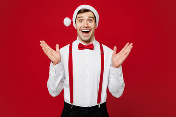 Poster - Merry young surprised fun shocked man wear white shirt Santa hat posing look camera spread hands isolated on plain red background studio portrait. Happy New Year Christmas celebration holiday concept