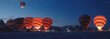 A panorama of the platform for preparing hot air balloons for takeoff, early in the morning, with hustle and bustle around them. Some balloons are taking off, igniting the fire inside.