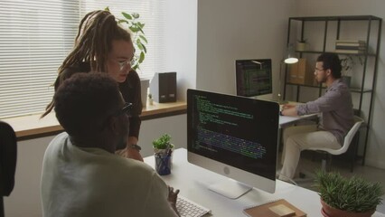 Medium shot of two diverse programmers having discussion of software program code on black computer screen while coworking in office at daytime