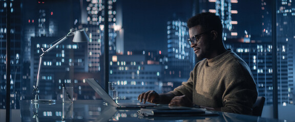 successful handsome black businessman working on laptop computer in big city office in the evening. 