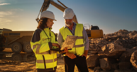 Poster - Caucasian Male Engineer And Hispanic Female Building Architect Walking On Construction Site With Laptop Computer And Talking About New Real Estate Project. Heavy Machinery Working. Golden Hour